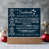 Navy blue sign with white text and floral designs on a wooden stand, set against a blurred decorative background.
